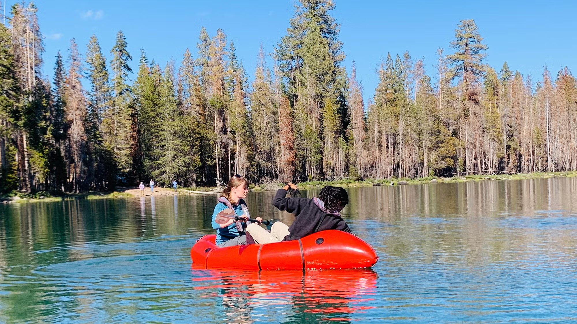 Two people in a red inflatable boat on a calm, sunny forest lake