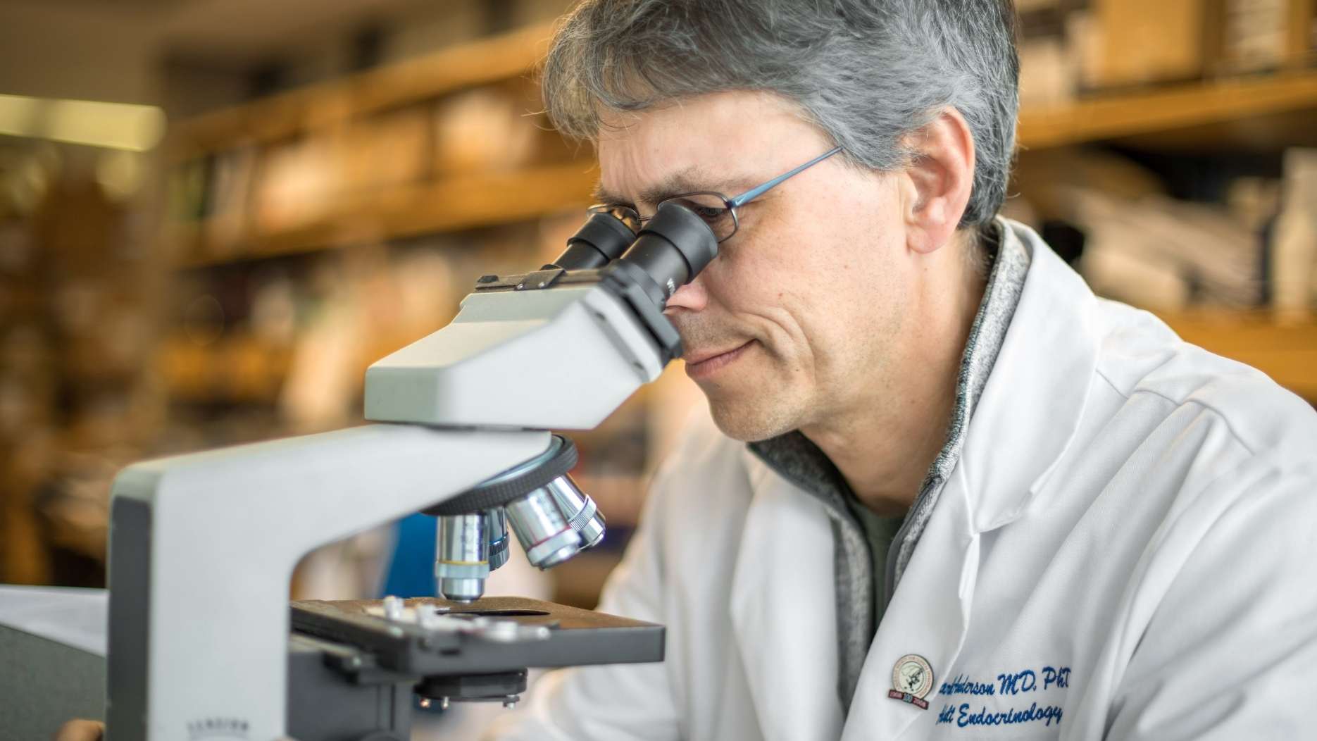 A lab scientist in a white coat looks through a microscope