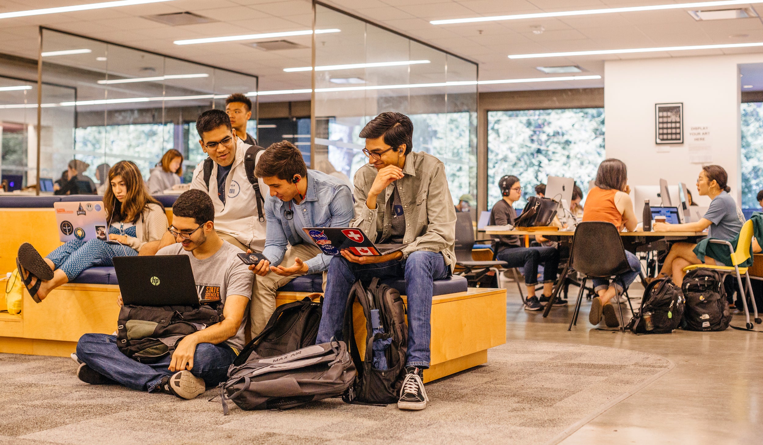 A group of college students work together in a modern campus study area. Some are sitting on benches and the floor with laptops and backpacks, while others collaborate at tables with desktop computers in the background.