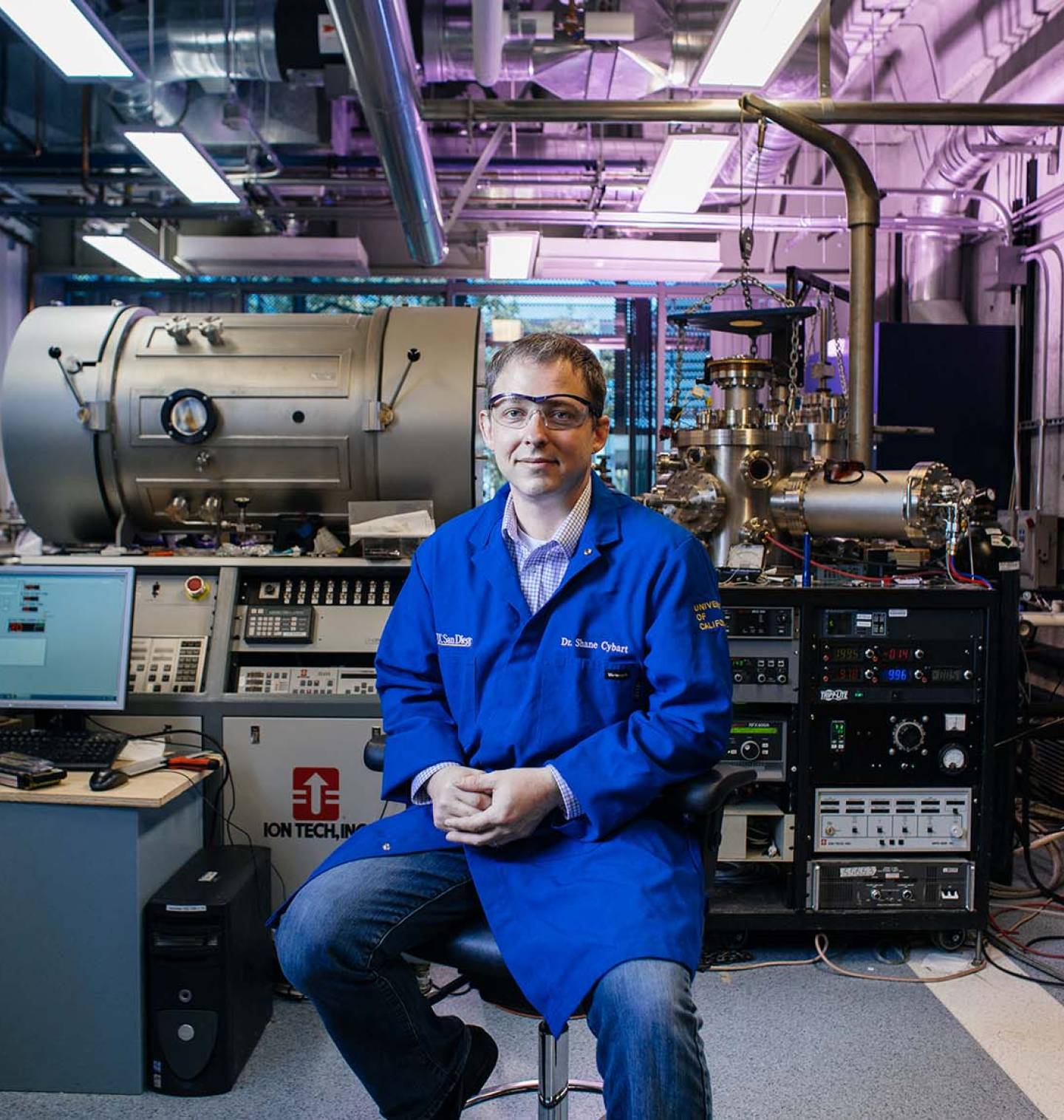 Wearing a blue lab coat, Dr. Shane Cybart sits on a stool in the Oxide Nano Electronics Lab at UC Riverside