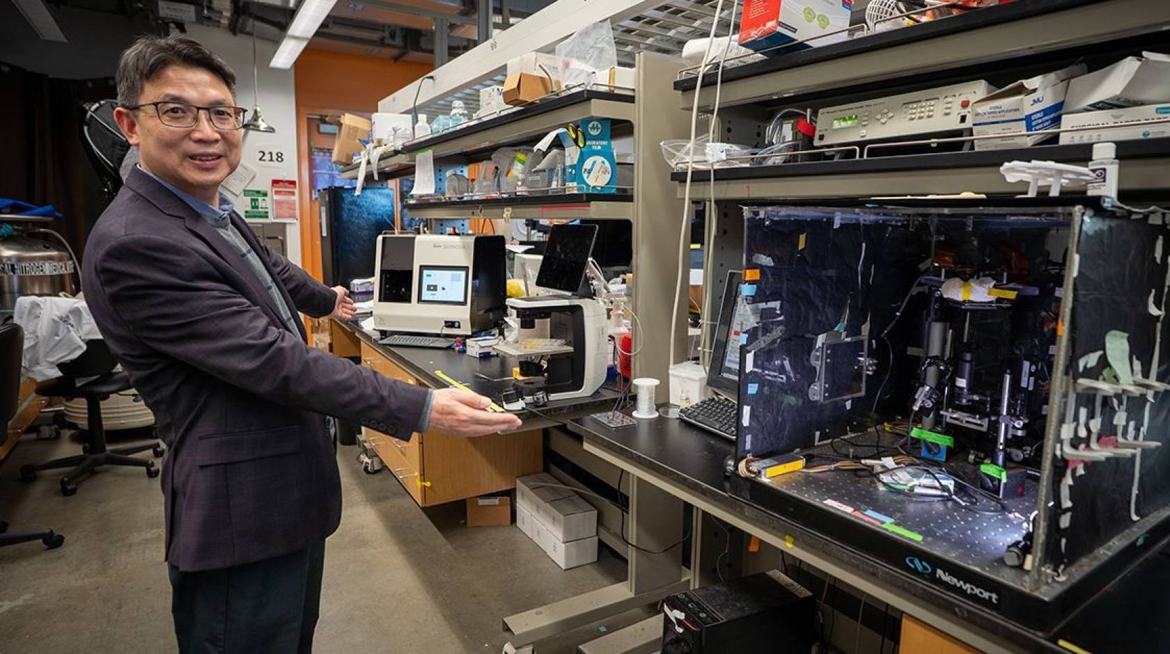 Man in a dark gray suit stands in a lab in front of multiple machines on a benchtop