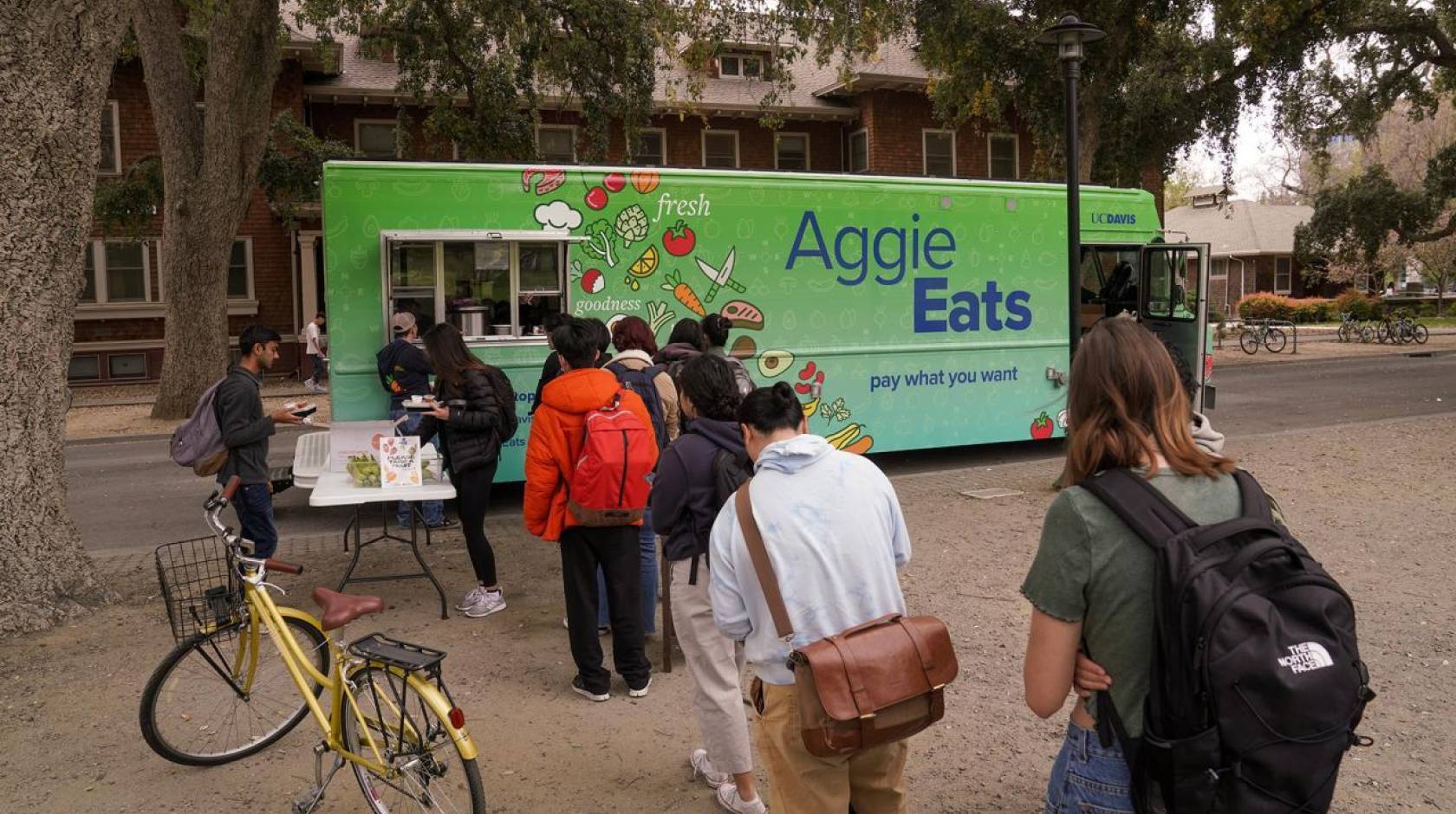 Students with backpacks and bikes line up in front of a food truck. The side of the food truck reads "Aggie Eats. Pay what you want."