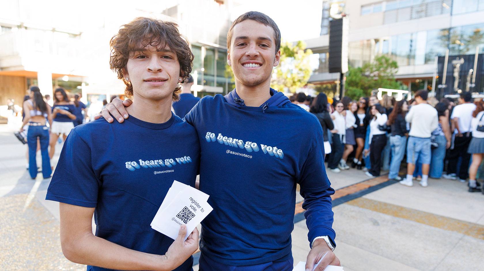 Two students smile for the camera with arms around each other's shoulders, wearing matching t-shirts that read "Go Bears Go Vote." One student holds a stack of printed QR codes labeled "Register to Vote"