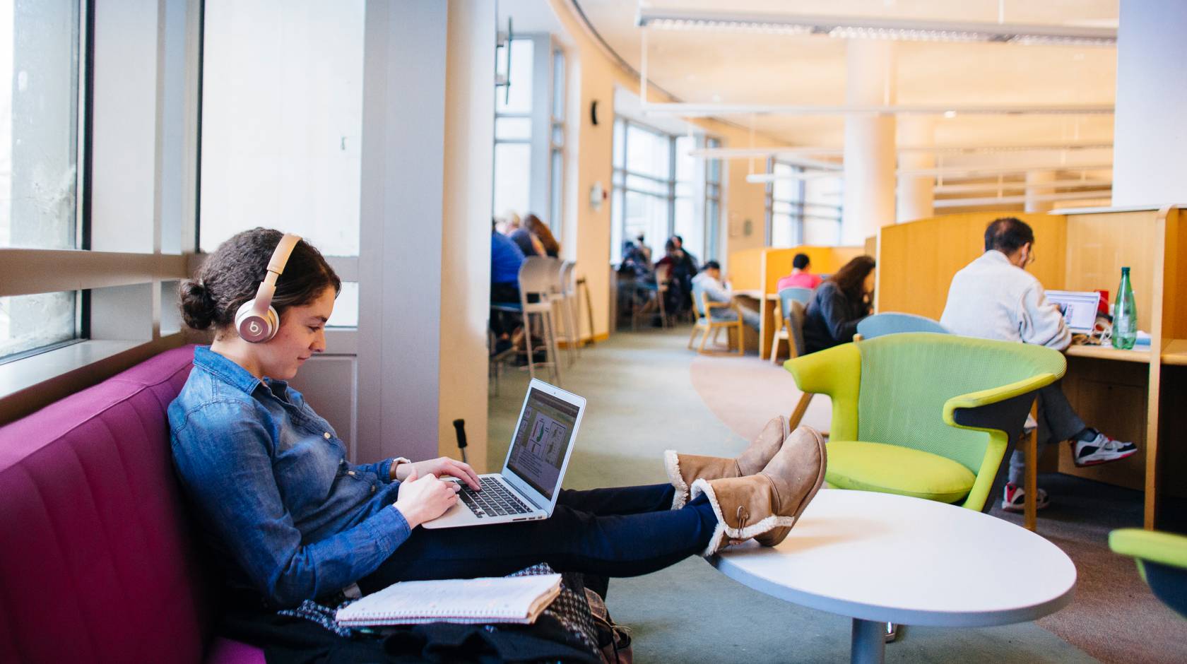 A woman sits on a sofa in a library typing on her laptop with her feet up in a table and headphones on.  Beyond her are more people studying at desks.