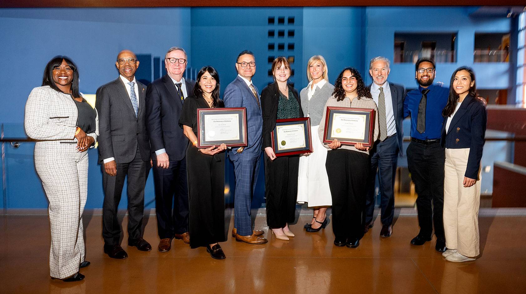 About ten people pose for a photo wearing business attire, three holding plaques.