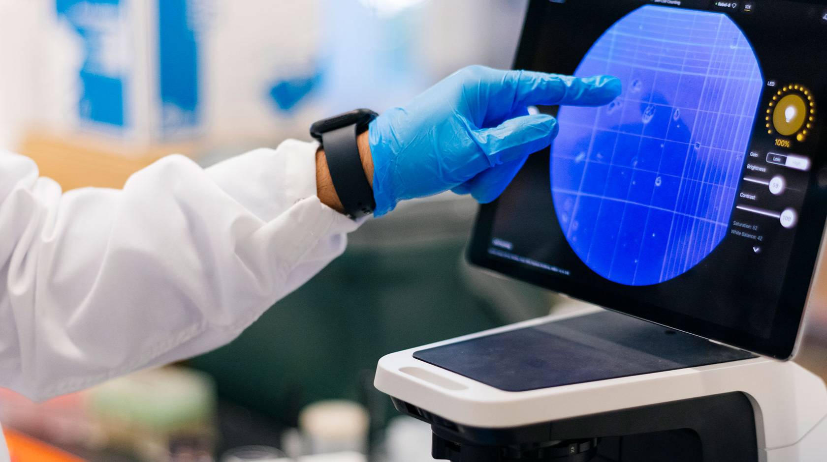 A hand in a blue glove points to a tablet screen showing a microscope view.
