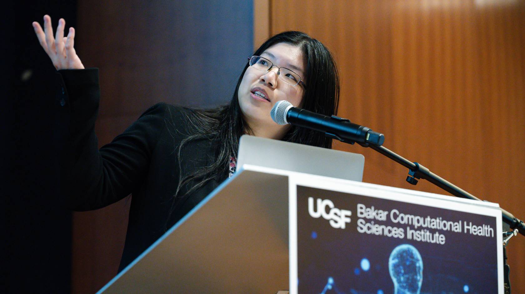 A young woman points at a screen off-camera while at a UCSF lectern