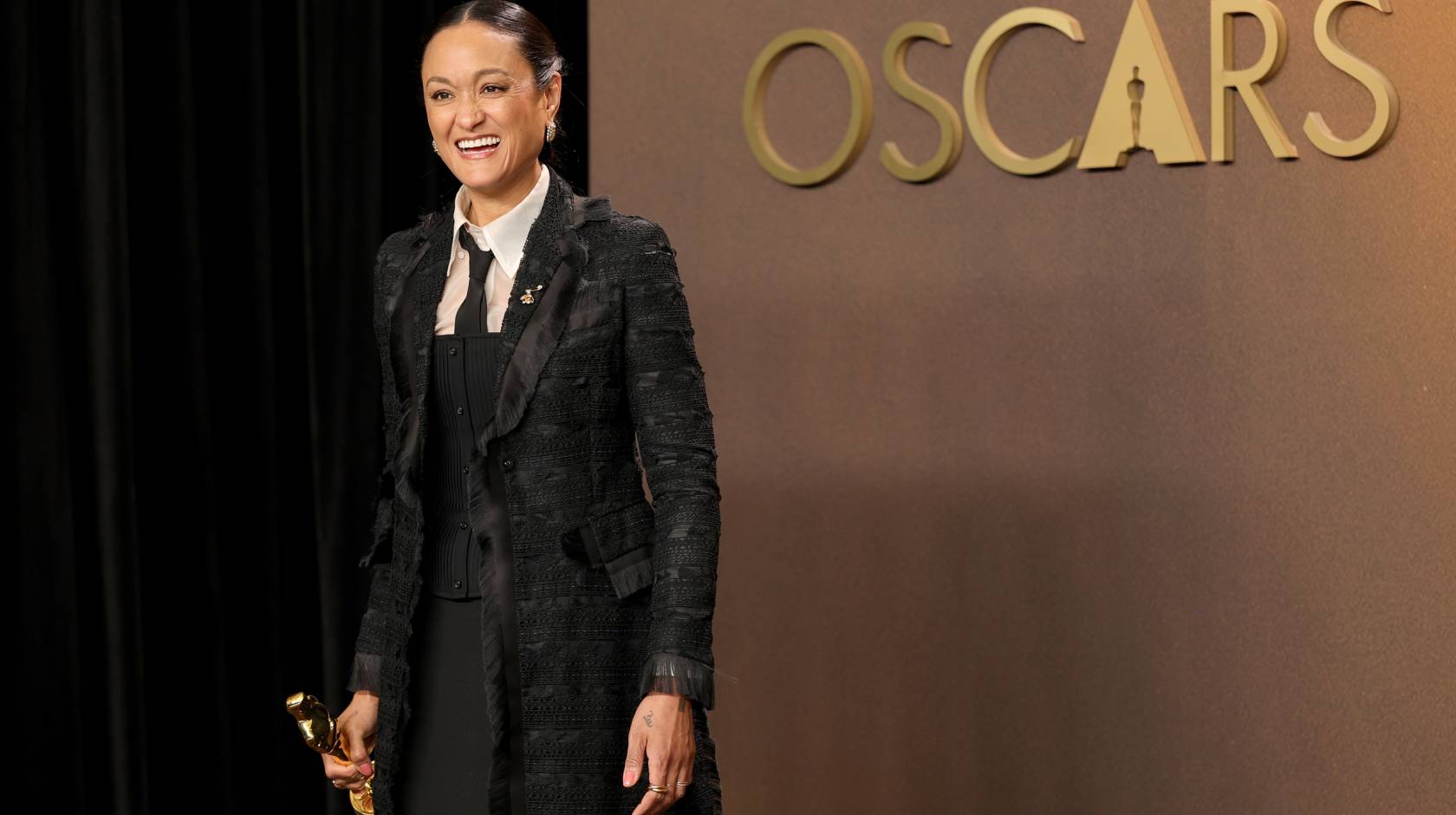 A woman in a black coat holds an Oscar smiling overjoyed in front of a gold background that says Oscars