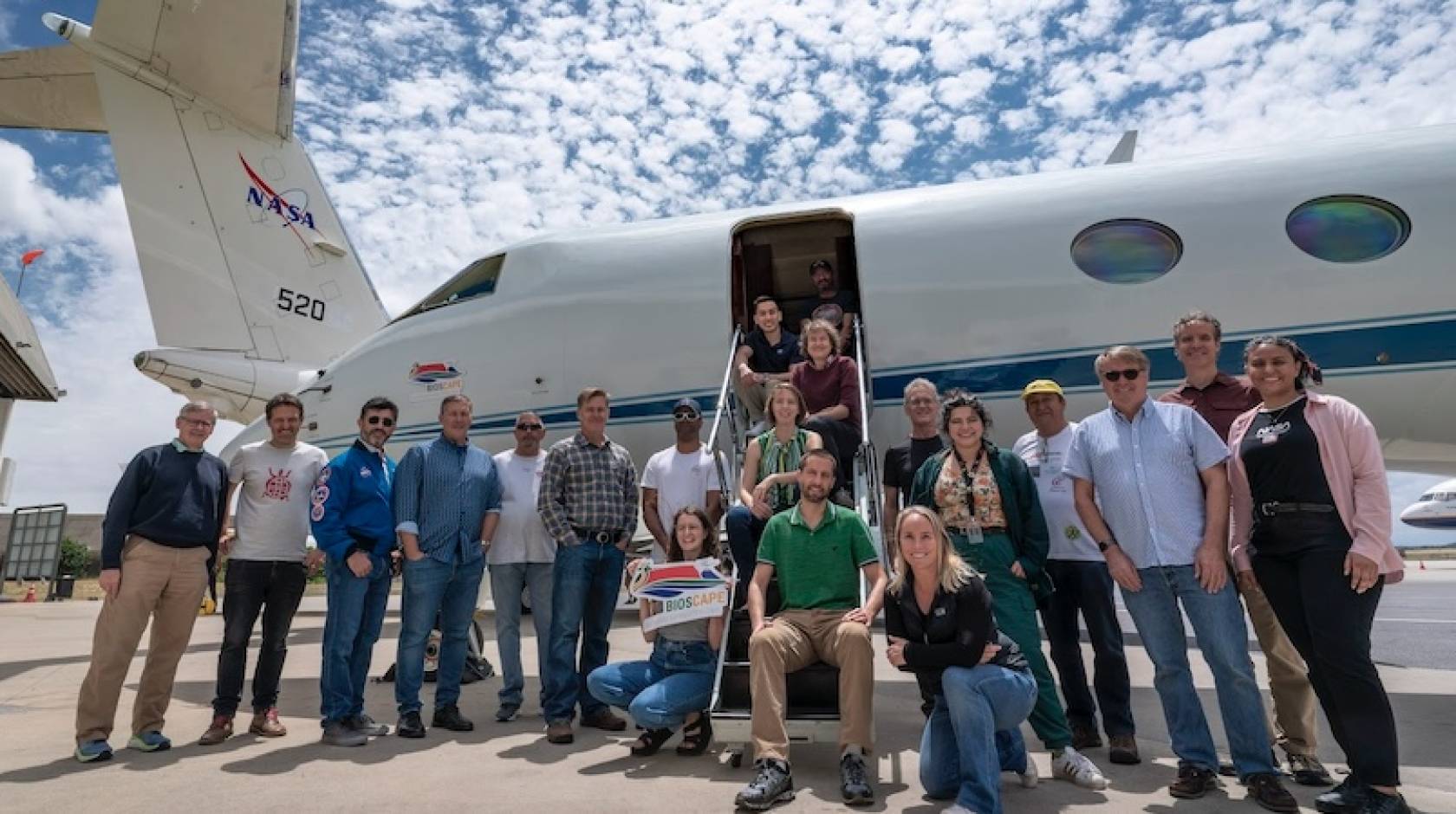 About 20 people gather for a photo on an airport tarmac, in front of a small jet