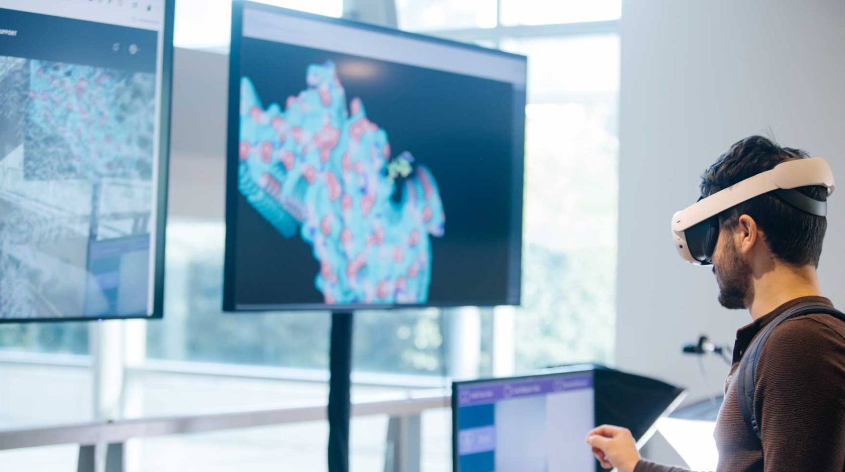 Someone wearing a virtual reality headset in front of two large screens showing molecules and one smaller screen