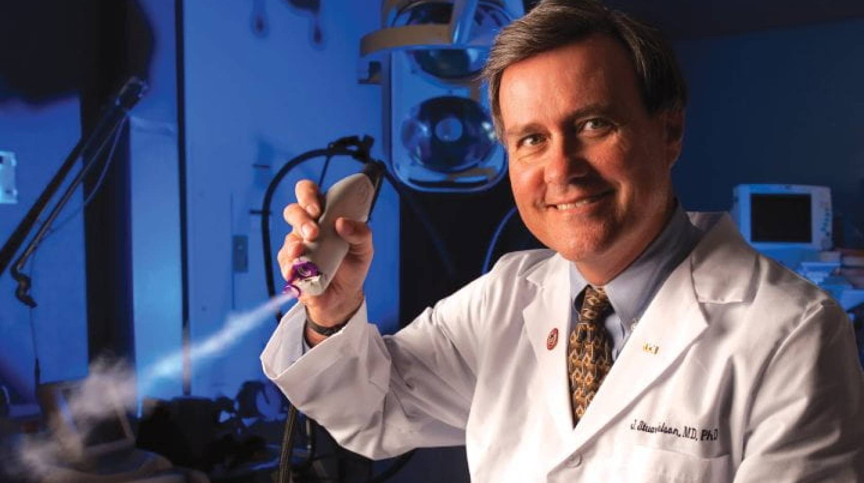 A surgeon in a white coat in a darkened lab holds a skin cooling device up to the camera and smiles.