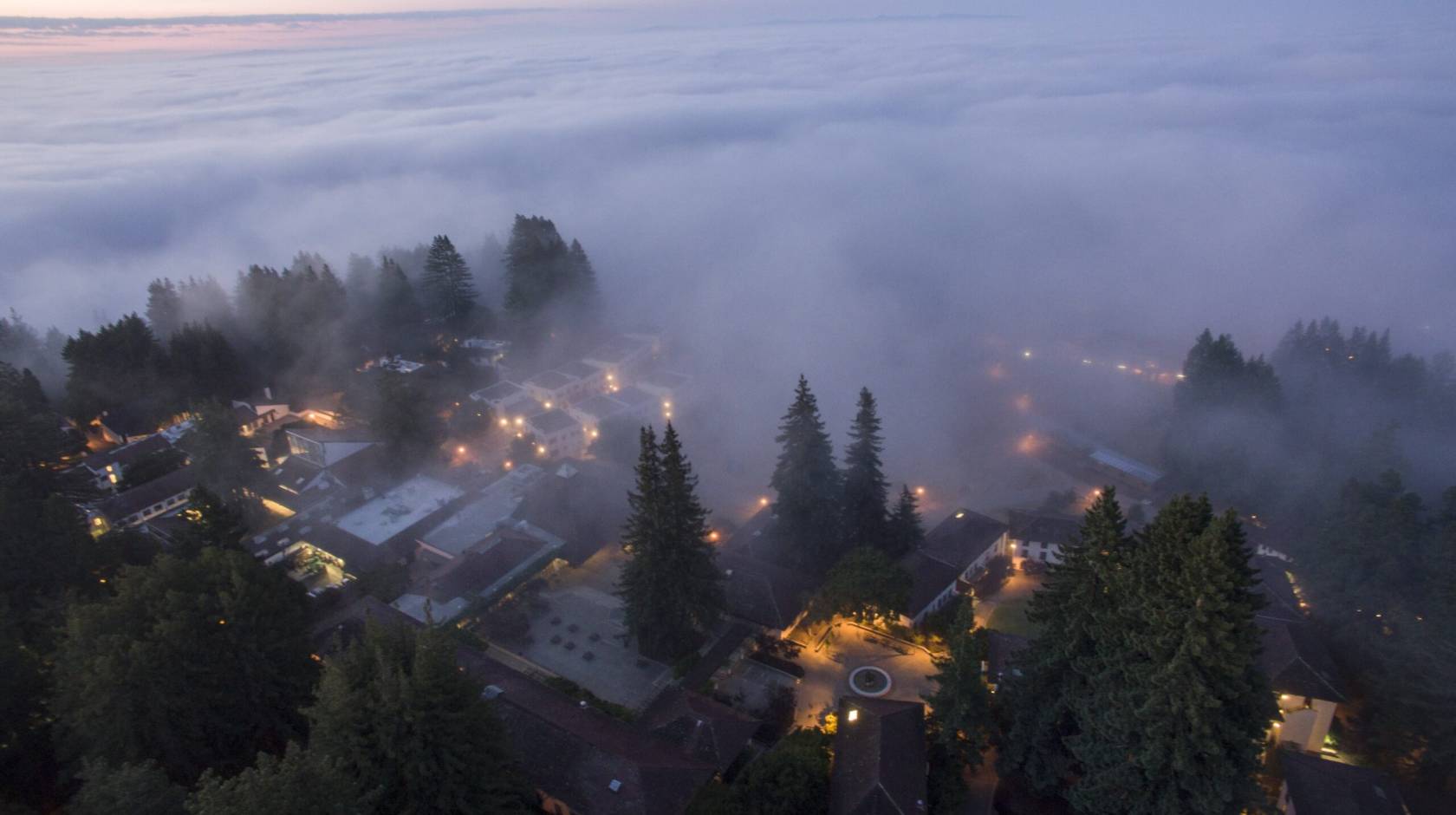 A view from above of heavy fog rolling from a reddish sky over a twilit UC Santa Cruz campus