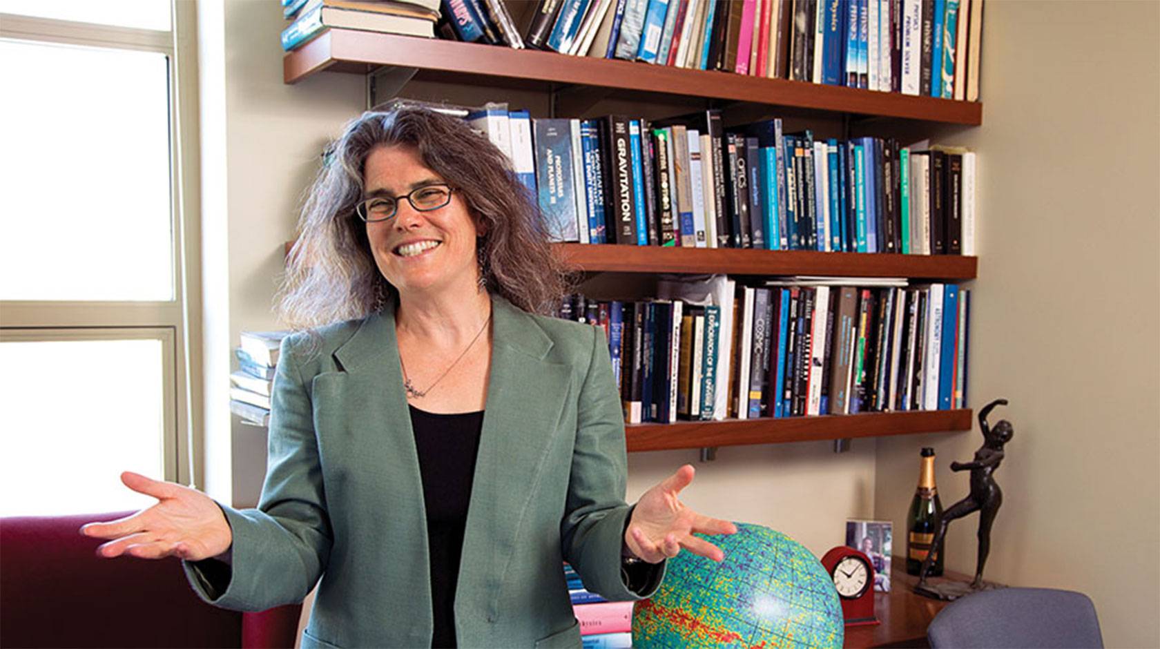 Andrea Ghez smiles and gesticulates with her hands, standing in front of a bookshelf and a globe