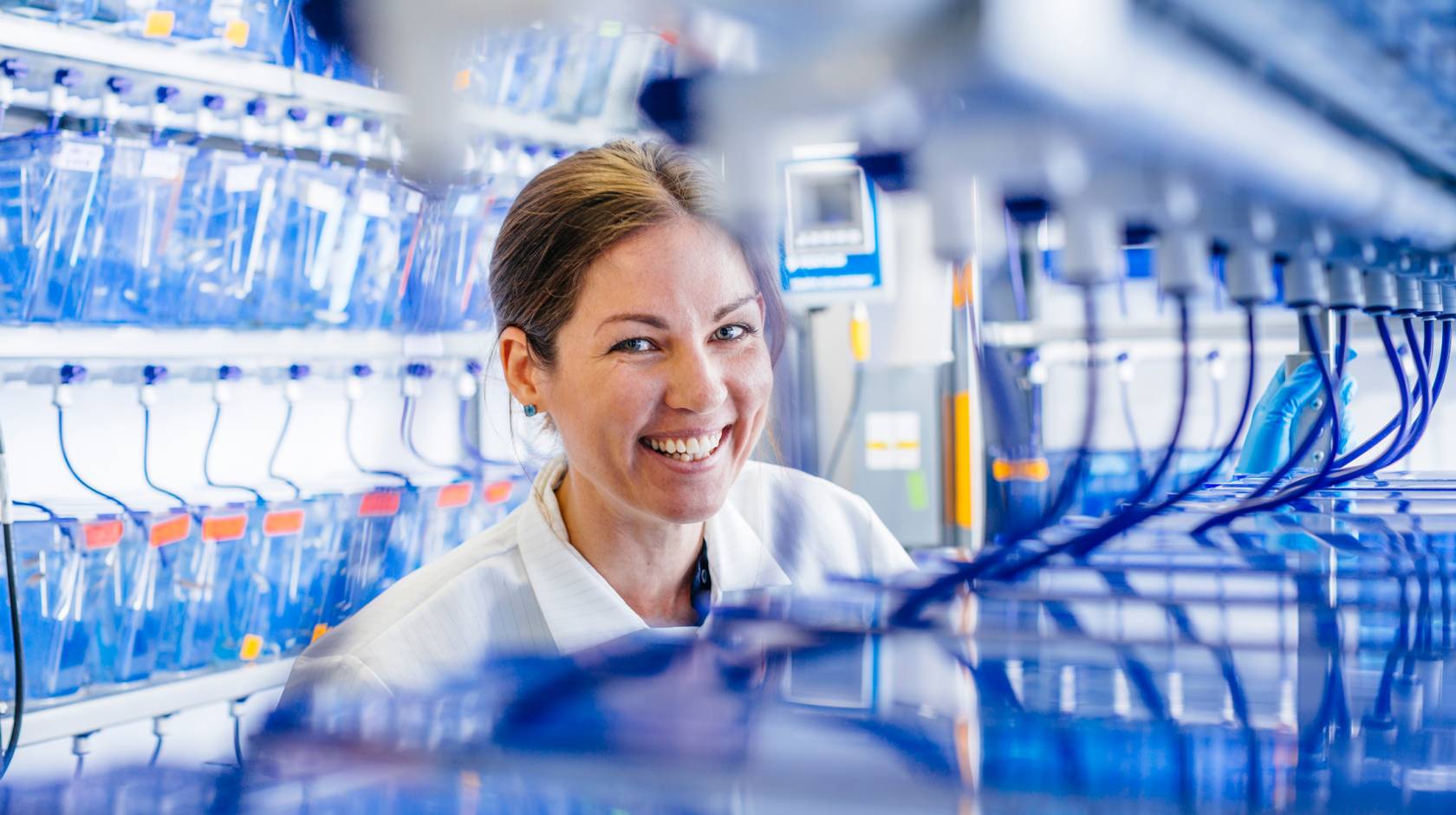 A student researcher smiles in a lab coat around blue tubing