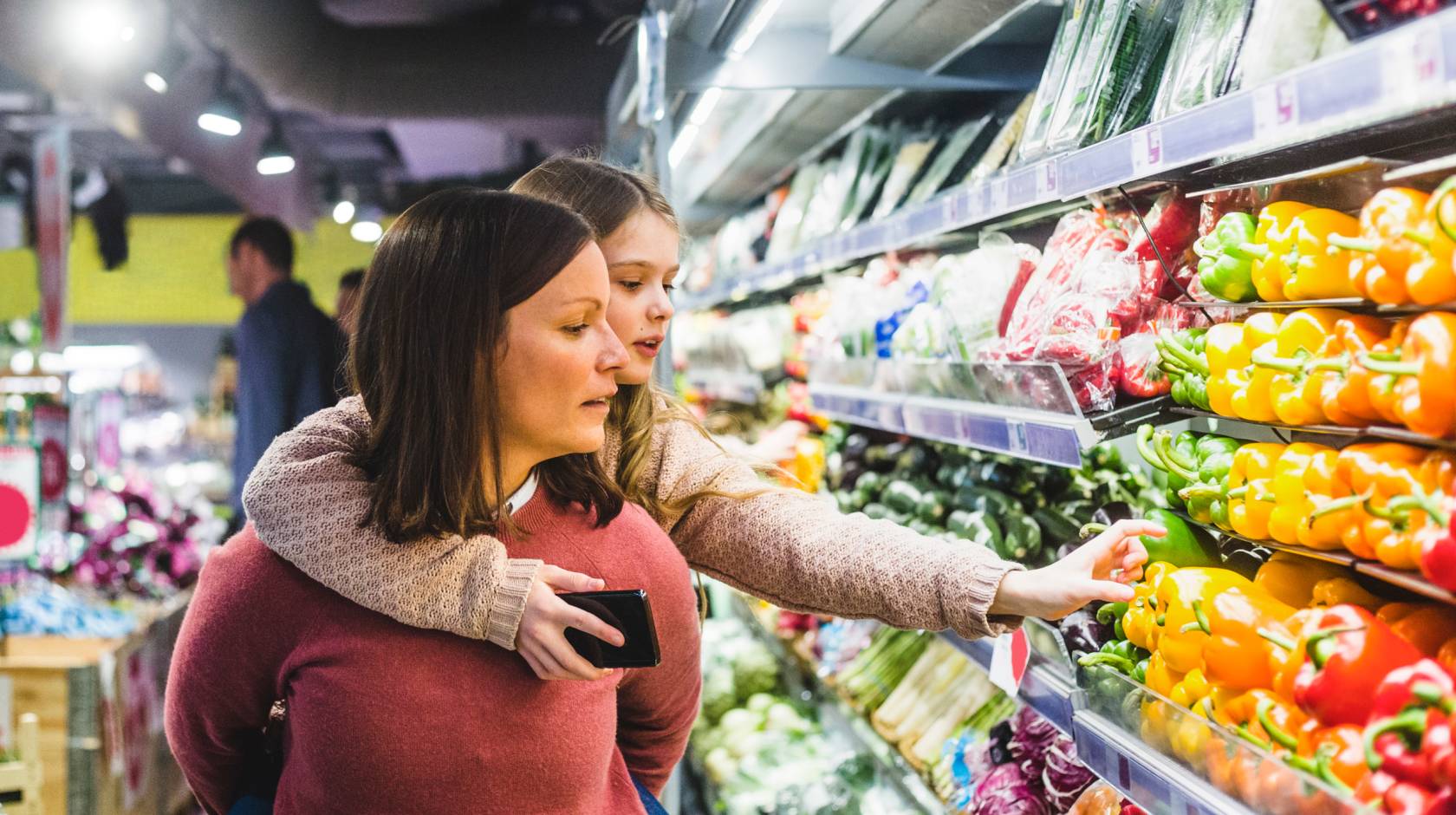 A child hanging on to her mom's back reaches for a pepper in the produce aisle