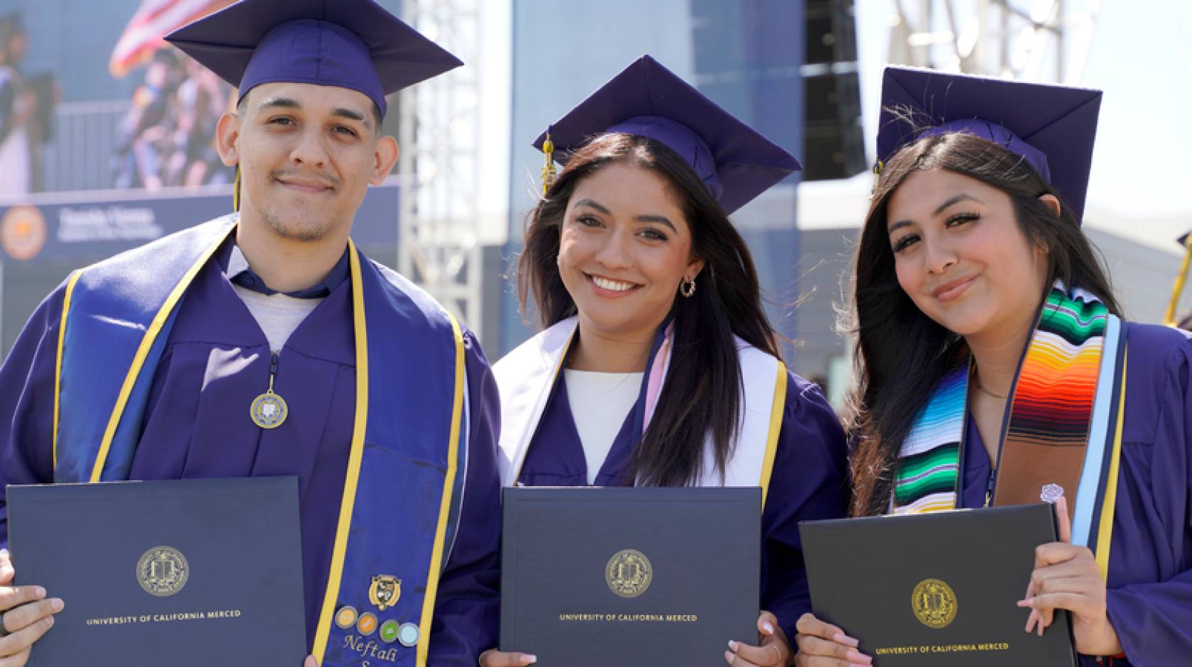 Three graduates in blue caps and gowns smile while holding their diplomas during a University of California, Merced commencement ceremony. They stand side by side, wearing stoles and tassels, with a stage and other graduates visible in the background.