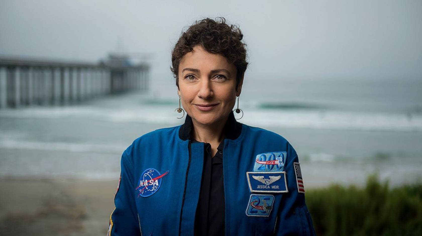 A woman in a blue NASA jumpsuit photographed with a pier and ocean behind