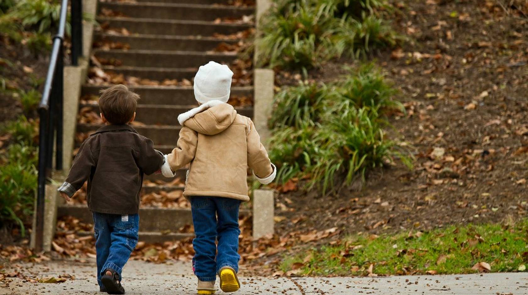 Two young children in winter clothes hold hands as they approach a park stairway, photographed from behind