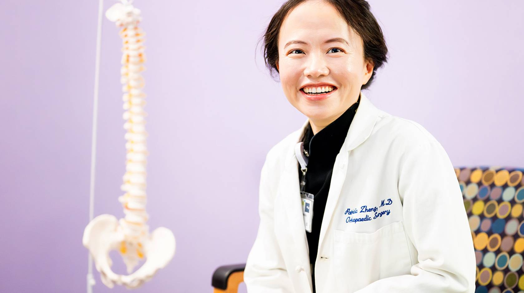 A woman with short hair in a white doctor's coat smiles in a chair with a model of a human spine behind her against a lilac backdrop