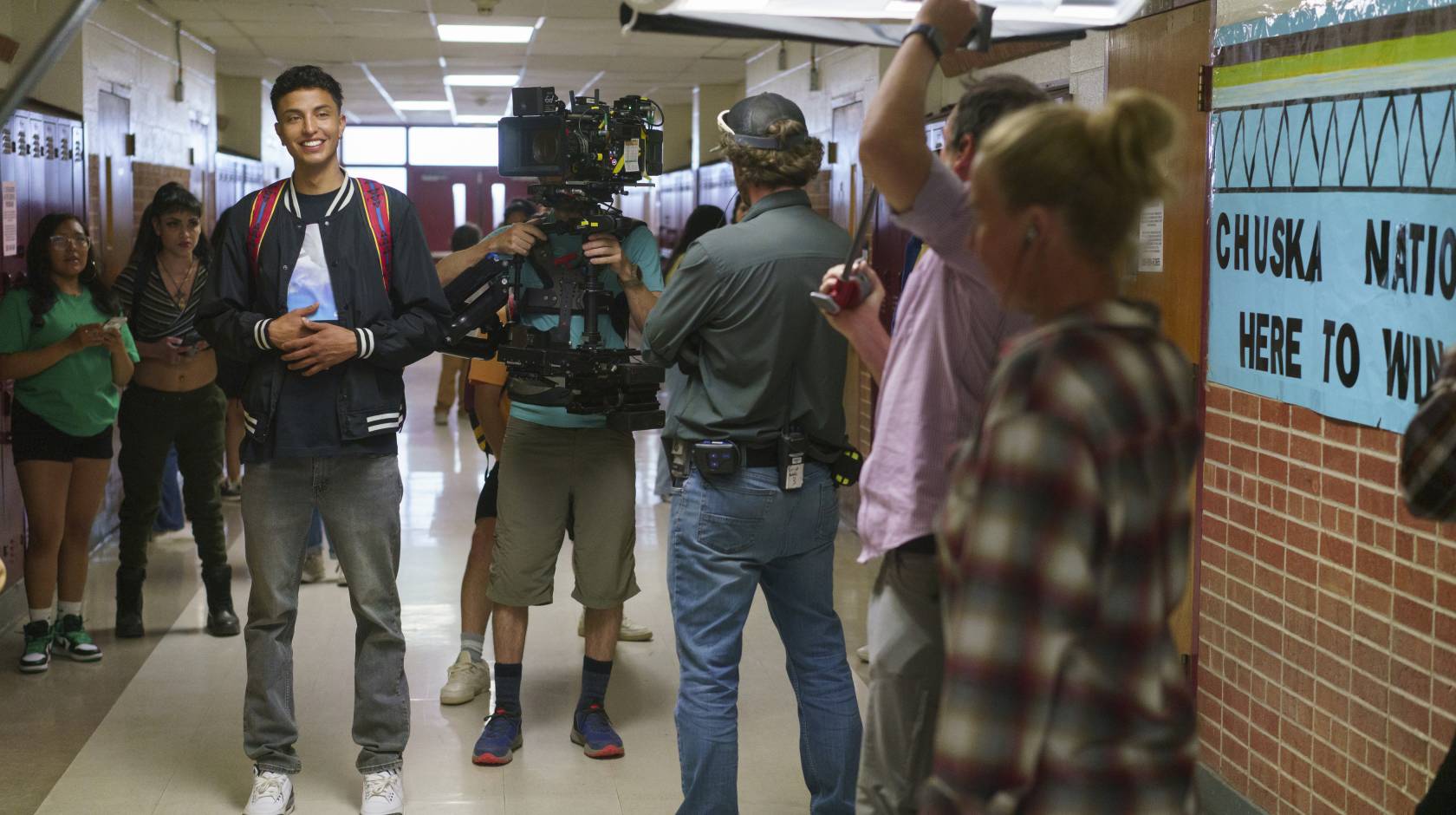 A young man smiling in a busy high school hallway being filmed as part of the movie Rez Ball