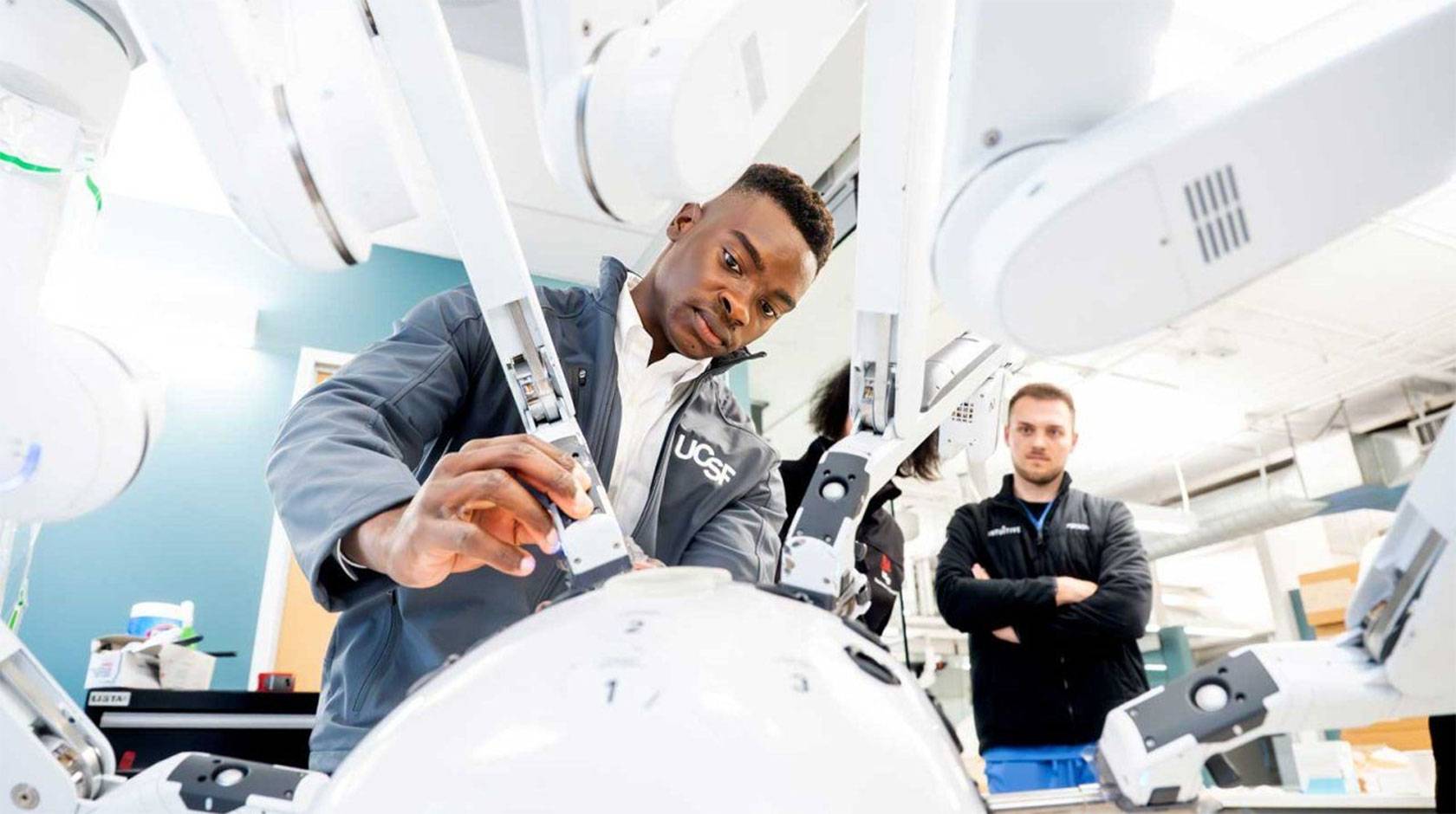 A student focuses intently as he operates one arm of a many-armed robot, while a teacher watches