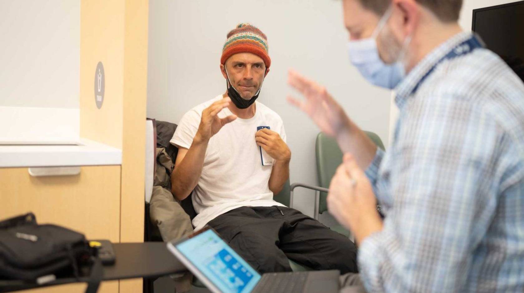 In the background, in focus, a man in a beanie makes a gesture with his hand as if to grasp. In the foreground, a doctor in a mask in front of a computer screen makes the same gesture