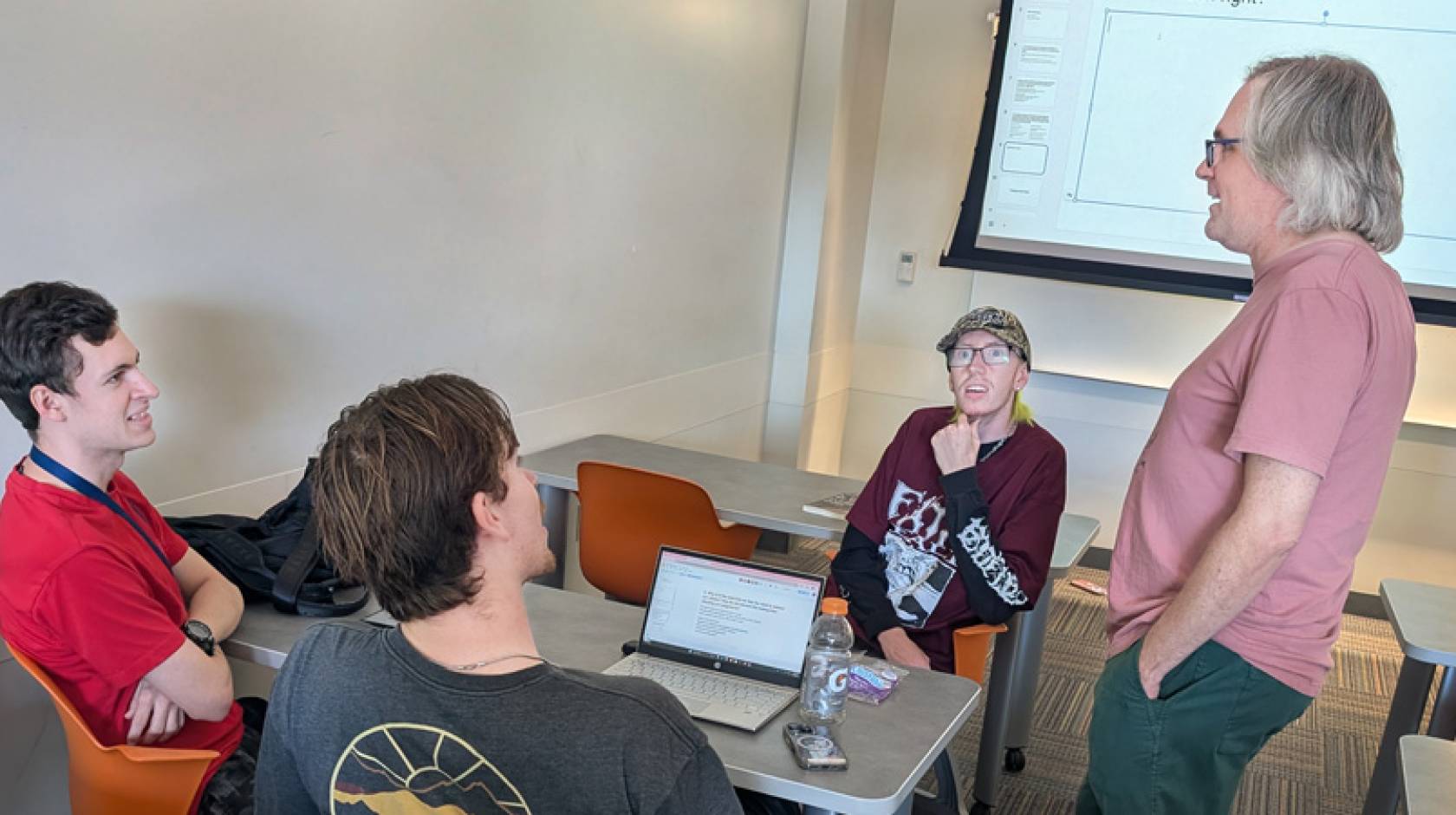 Three seated people talk to a standing professor in a classroom