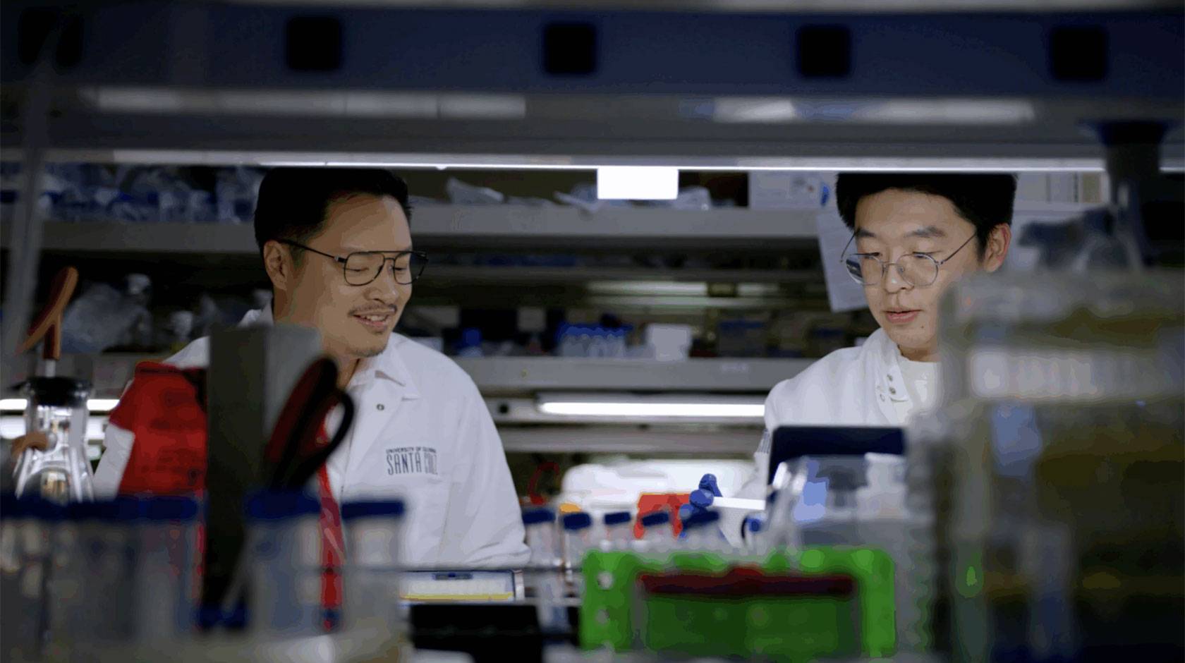Two scientists in white coats work at a lab bench