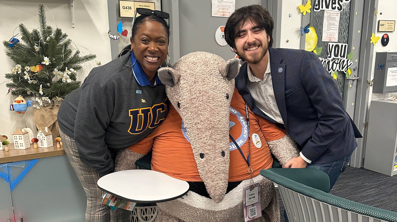 In a student center space, on the left, a woman with a sweatshirt that says UC and on the right, a young man with a beard smiling; center, a stuffed Anteater, the UC Irvine mascot