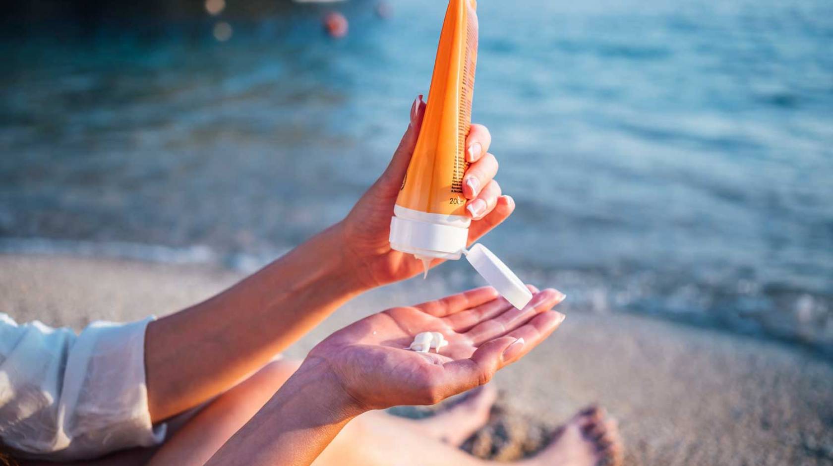 Someone sitting on the beach holds a tube of sunscreen in one hand with the other with a dab of sunscreen on it