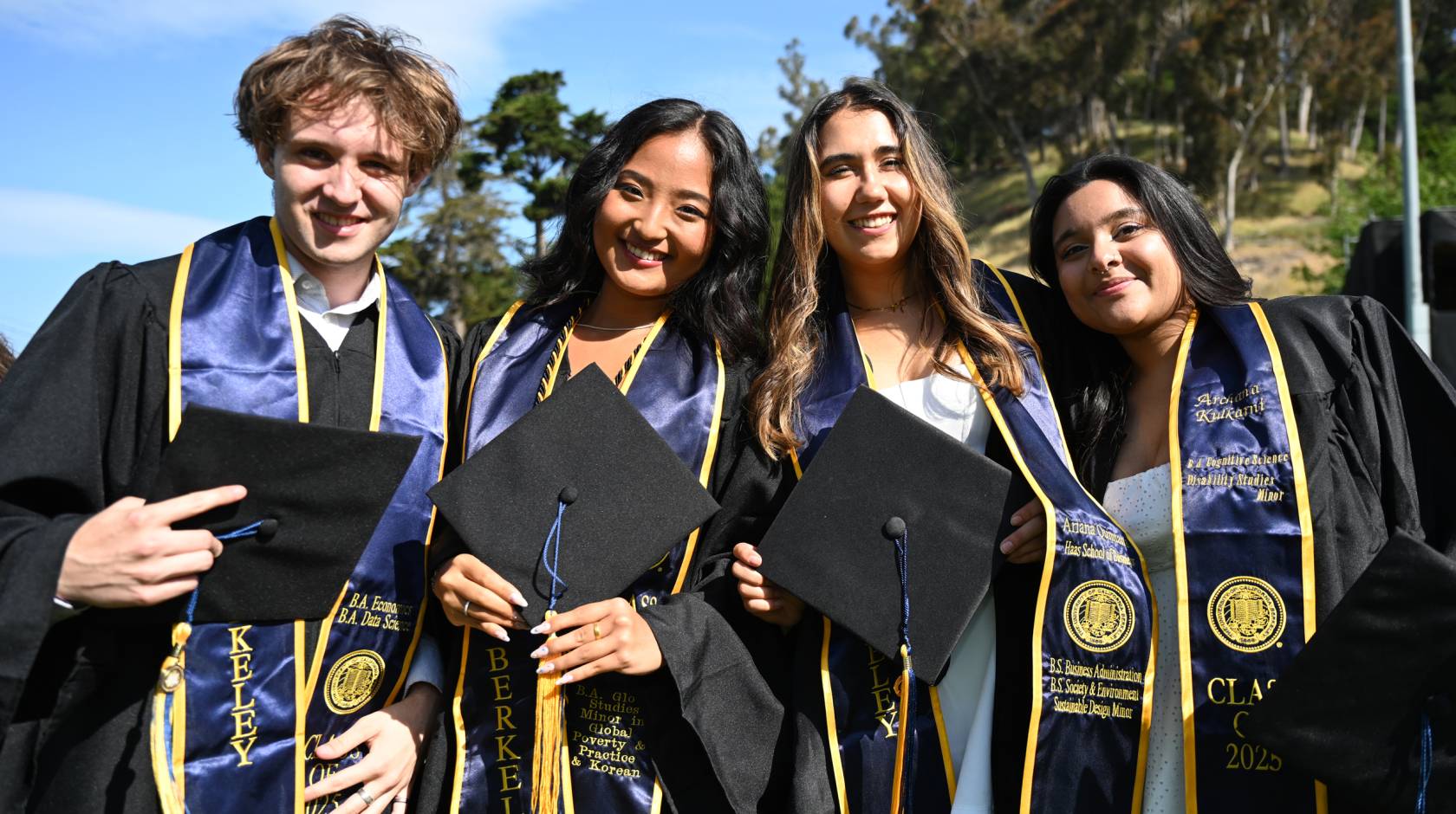 Four graduates in robes and stoles hold their caps and smile at the camera