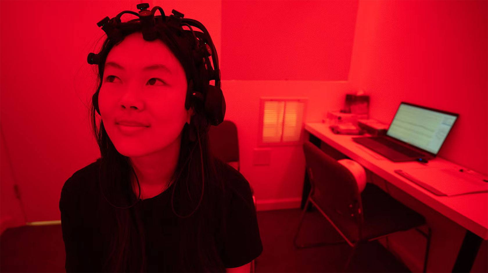 A woman wearing a device on her head sits in a lab bathed in deep red light