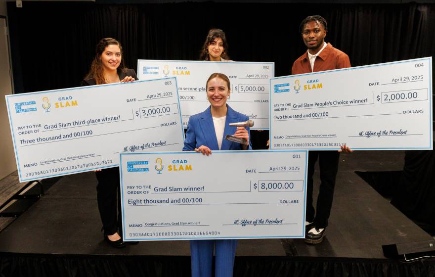 Four people, one in the foreground, three in the background, hold up giant checks