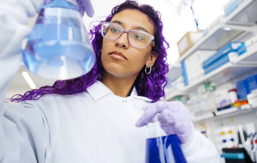 Woman with long, dyed-purple hair wearing a white lab coat and protective gear compares two flasks full of colorful liquid in a lab