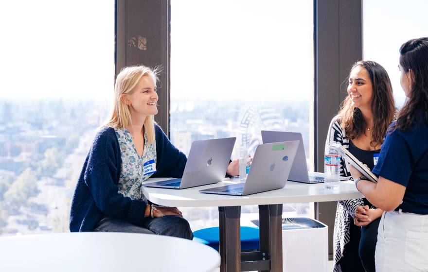 Three women sit with laptops at a table in front of a wall of windows