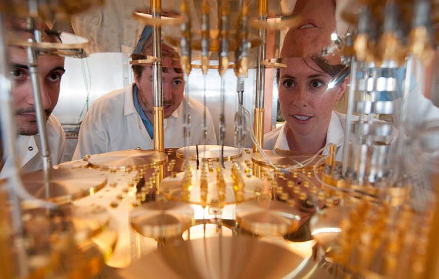 Three people in white lab coats peer into a very large, delicate, gold-colored machine.