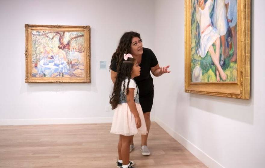 A mother and her young daughter talking about a painting they are looking in a museum gallery.