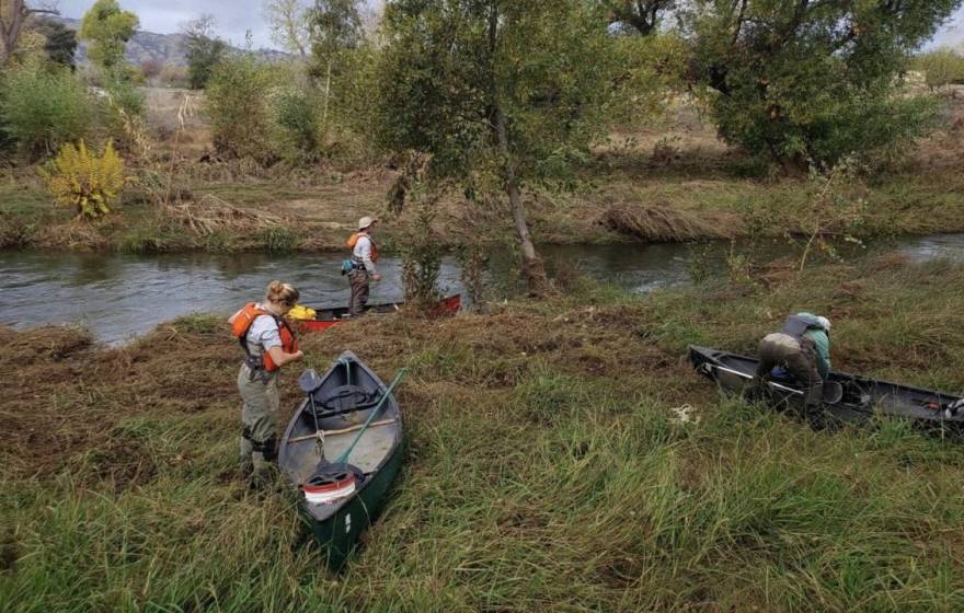 Three people with water gear and canoes on the bank of a creek
