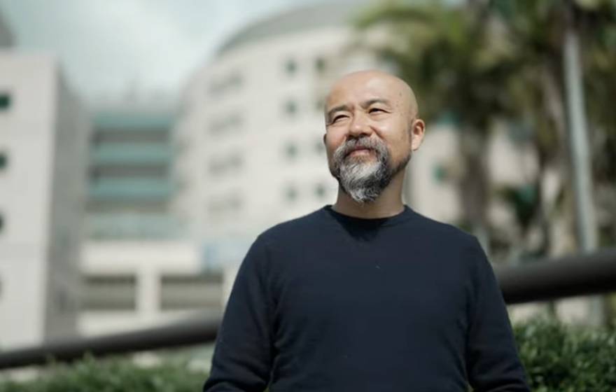 Hirotaka Matsunaga looks pensively beside the camera in front of a medical building