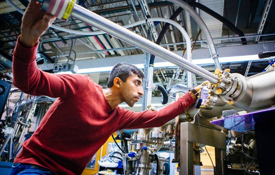 A grad student focuses intently as he slides a three-foot-long steel bar into a large apparatus made of tubes and wires, in a lab full of similar such apparatuses. 