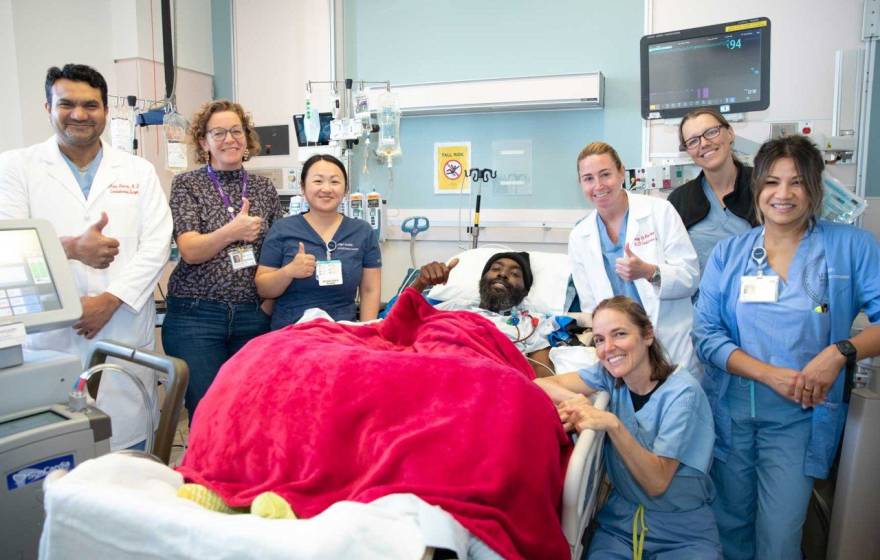 A man in a hospital bed with a red blanket on him is surrounded by a team of nurses and doctors, smiling and giving thumbs up
