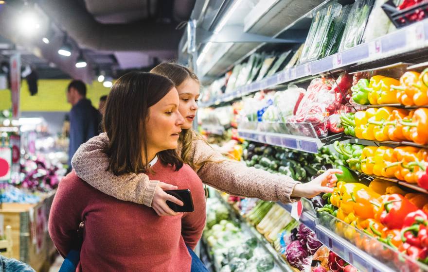 A child hanging on to her mom's back reaches for a pepper in the produce aisle