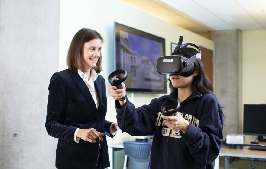 A woman in a jacket and blouse smiles next to someone in a VR headset with a sweatshirt