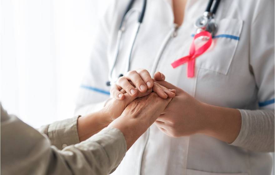 A patient, left, and medical care provider holding hands; provider has a pink breast cancer ribbon on her lapel