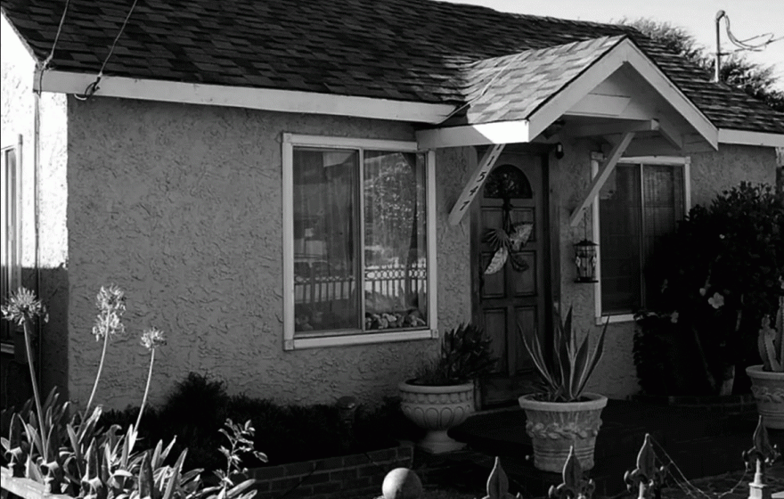 Black and white photo of a modest stucco house with eerie green lights flashing in the window