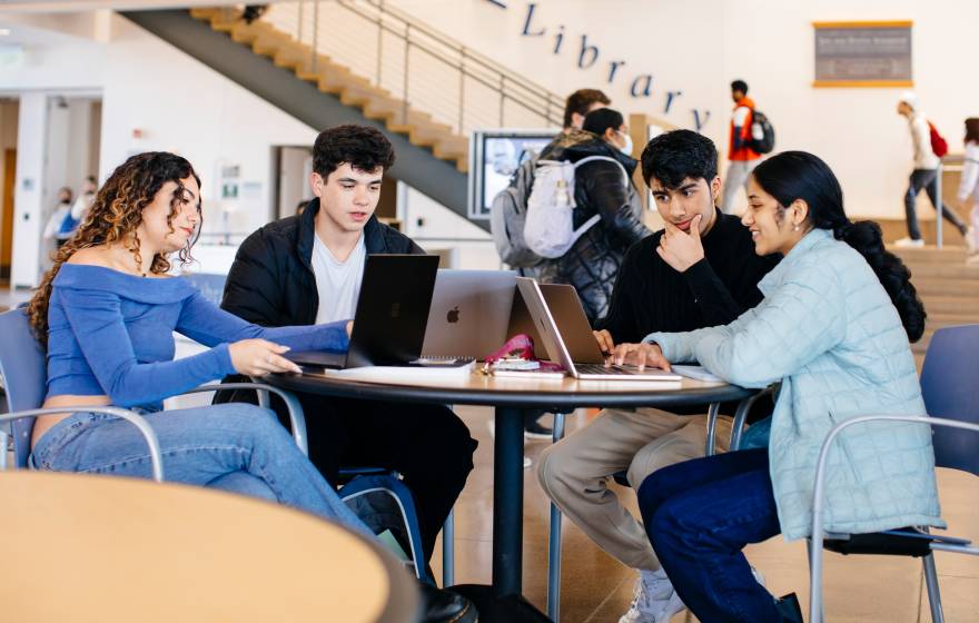 A group of students studying together at a table in a busy campus space, with a sign that says "Library" in the background. 