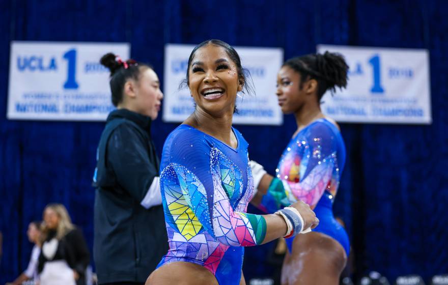 Jordan Chiles smiles while at a competition in a leotard, with another gymnast behind her on her right and a trainer behind her on her left