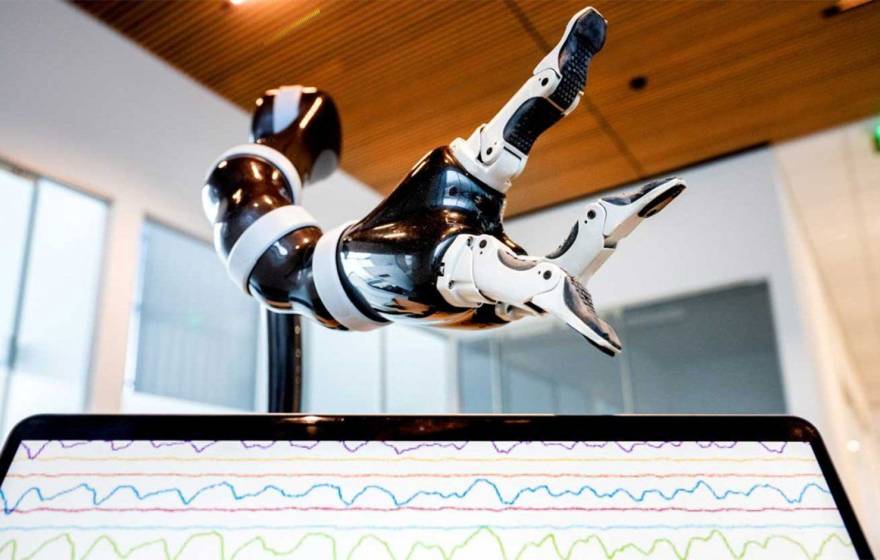 A robotic arm hovers over a computer screen showing brainwaves as a stack of tiny lines. 