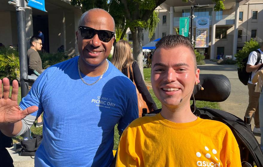 Ryan Manriquez, who uses a motorized wheelchair and wears a yellow shirt with the logo of the Associated Students of UC Davis on it, smiles for a photo alongside UC Davis Chancellor Gary May, who wears sunglasses and a blue t-shirt and is waving at the camera