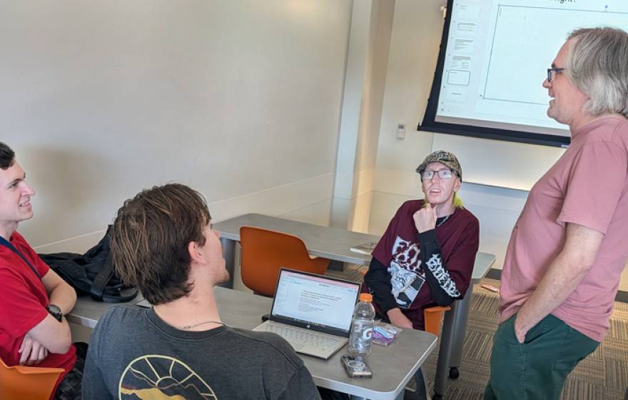 Three seated people talk to a standing professor in a classroom